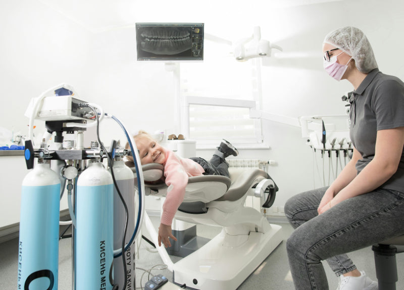 Young child reclines comfortably in a dental chair, smiling at a masked dental professional during a calm, child-friendly first dental visit.