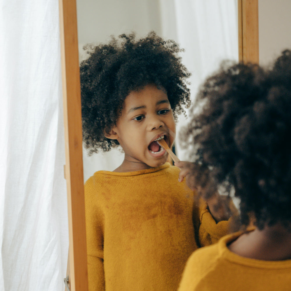 A young child brushes their teeth while looking into a mirror in a softly lit bathroom. The child’s focused expression shows learning and independence, highlighting how daily brushing routines support healthy smiles at home.