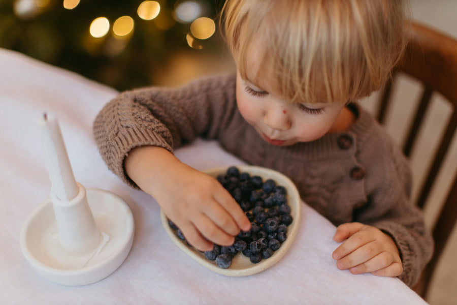 A small child seated at a table picks blueberries from a small bowl, leaning in with focus. A white candle holder sits nearby, and soft warm lights blur in the background.