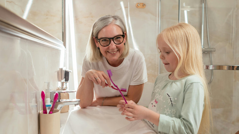 Parent and child smiling at sink while brushing together, showing how to help your child brush their teeth gently