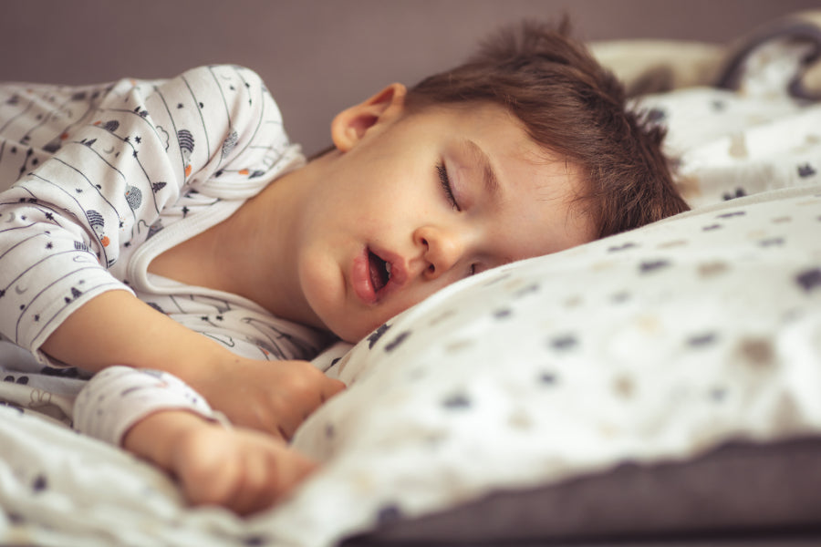A young child sleeps on their side with their mouth slightly open, resting on a patterned pillow in soft natural light. Example of mouth breathing in children.