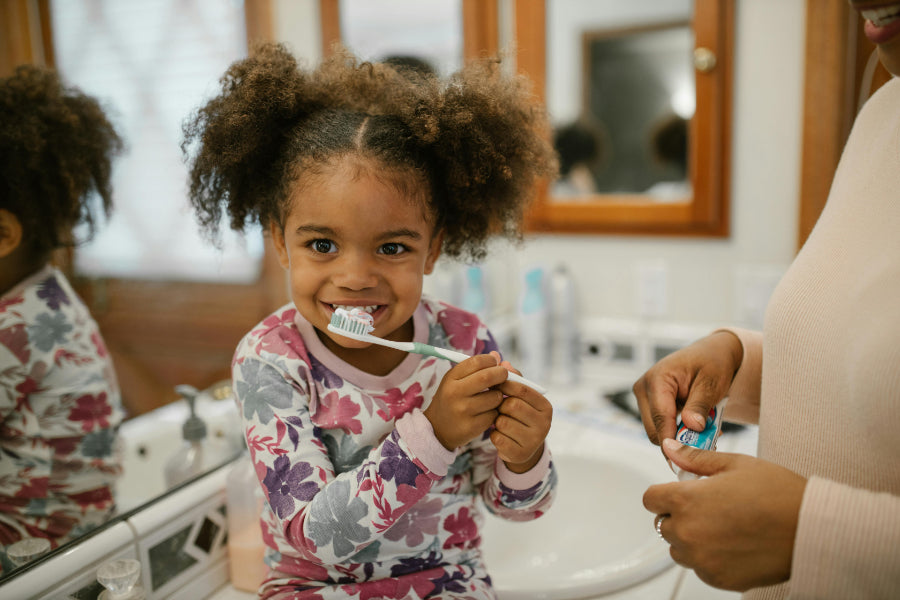 A young girl with curly hair, wearing floral pajamas, smiles while brushing her teeth in a bathroom. An adult's hands are visible on the right, holding a tube of toothpaste near the sink. | reinspire your child's toothbrushing routine this winter